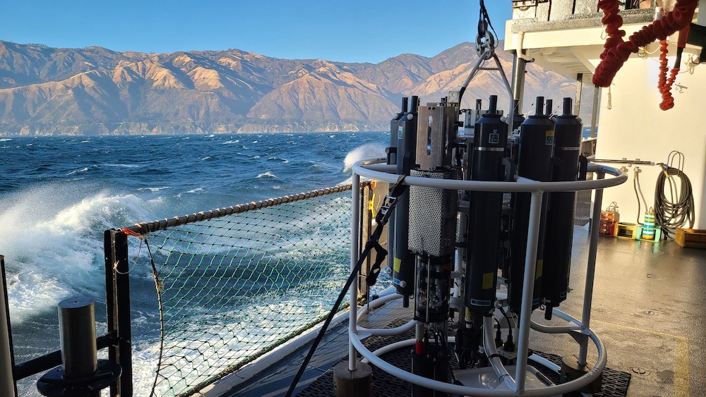 A round oceanographic sampling instrument with thin vertical tanks surrounded by a pipe frame attached to a cable sits on the deck of a ship sailing to the right a good ways from a mountainous shore.